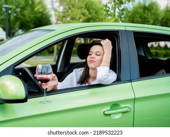 Beautiful Young Modern Girl Driving A Car With A Glass Of Red Wine In Her Hand. The Female Driver Is Alone In The Car, Drinking Wine. Drunken Stalker. Violation Of Traffic Rules