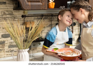 Beautiful Young Mom Cook In An Apron With Her Son Prepares Cookie Dough In A Wooden Bowl At Home In The Kitchen. Selective Focus. Portrait. Close-up