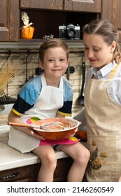 Beautiful Young Mom Cook In An Apron With Her Son Prepares Cookie Dough In A Wooden Bowl At Home In The Kitchen. Selective Focus. Portrait. Close-up