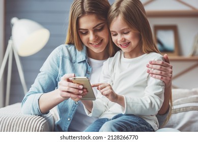 Beautiful young mom and her little daughter are using a smartphone and smiling while sitting on sofa at home - Powered by Shutterstock