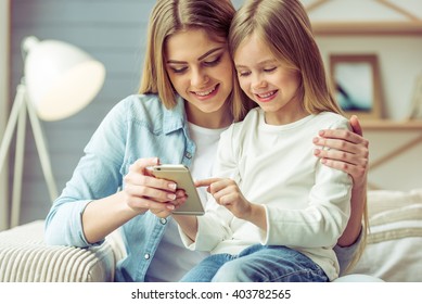 Beautiful young mom and her little daughter are using a smartphone and smiling while sitting on sofa at home