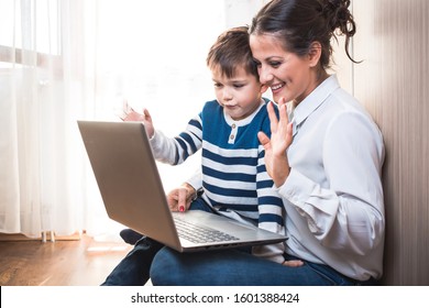 Beautiful Young Mom And Her Son, Sitting On The Floor Looking At Laptop And Waving. Video Call With Someone