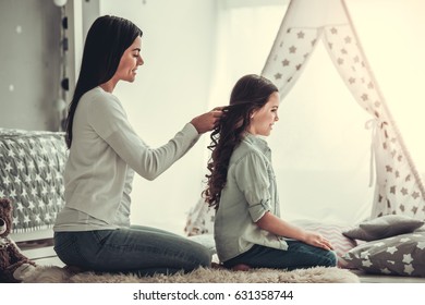 Beautiful young mom is making her cute daughter braids. Both are smiling while sitting in girl's room at home - Powered by Shutterstock