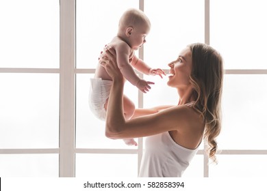 Beautiful Young Mom Is Playing With Her Cute Baby And Smiling While Standing Near The Window At Home