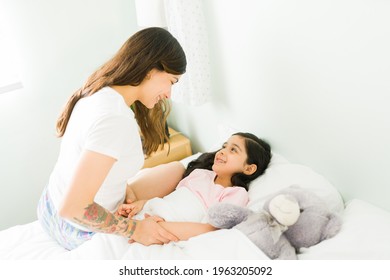 Beautiful young mom smiling to a little girl and tucking her in bed at night. Adorable daughter telling goodnight to her mother