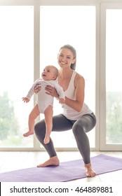 Beautiful young mom in sports wear is smiling while doing yoga with her charming little baby on a mat against window