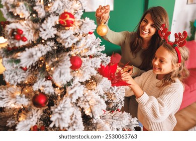 Beautiful young mother and cute daughter having fun decorating Christmas tree at home, placing Christmas lights and ornaments on it - Powered by Shutterstock