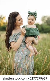 Beautiful Young Mother With Dark Hair And Green Dress Lifting Up Her Cute Little Daughter On Hands Playing Using A Blade Of Grass On Boho Picnic. Family Time Outdoors.