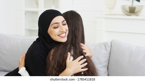 Beautiful Young Mother In Hijab Talking With Little Teen Daughter. Muslim Woman In Headscarf Having Talk With Little Girl On Couch At Home And Hugging. Mom's Love Hugs. Rear. Back View.