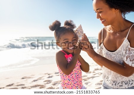 Beautiful young mother holding seashell near happy daughter ear at beach while she listening the sound of the ocean. Cute little child hearing to cockleshell with mom at tropical beach with copy space
