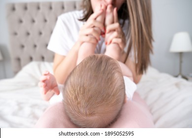 Beautiful young mother kisses her cutie baby girls's feet on bed. White background. Selective focus. Empty space for text.  - Shutterstock ID 1895052595
