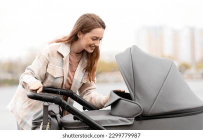 Beautiful, young mother pushing stroller with baby, looking at him, outdoors. Cheerful motherhood, care, parenthood, healthy lifestyle