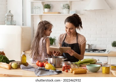 Beautiful Young Mother Reading To Adorable Daughter On Kitchen. Mom And Child Learning Recipe For Cooking Dinner. Happy Family Engaged In Food Preparation At Home. Girl Sit On Table Full Of Vegetable