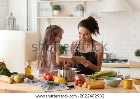 Beautiful young mother reading to adorable daughter on kitchen. Mom and child learning recipe for cooking dinner. Happy family engaged in food preparation at home. Girl sit on table full of vegetable