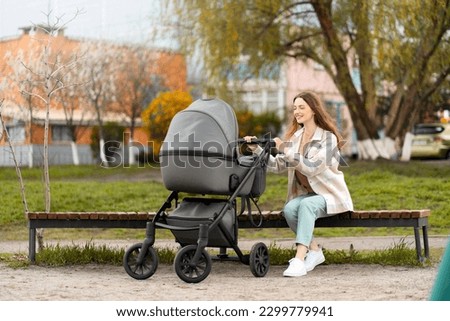 Beautiful, young mother sitting on bench, looking at stroller with baby, walking outdoors. Woman smiling at child, healthy lifestyle