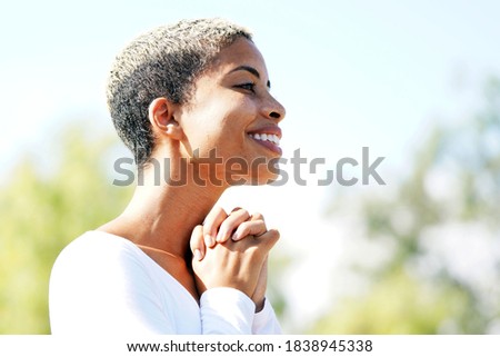 Beautiful young multicultural woman in the park on a clear, sunny day turned to the side for a closeup profile portrait while she clasps her hands in gratitude outside                               
