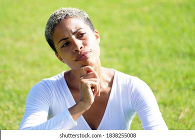 Beautiful Young Multicultural Woman Sits On A Grassy Lawn At A Park With White V Neck Shirt And Short Hair Thinking And Holding Hand To Chin With Furrowed Brow                           