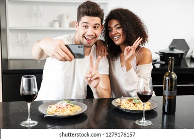 Beautiful Young Multiethnic Couple Having A Romantic Dinner At Home, Drinking Red Wine And Eating Pasta, Toasting, Taking A Selfie