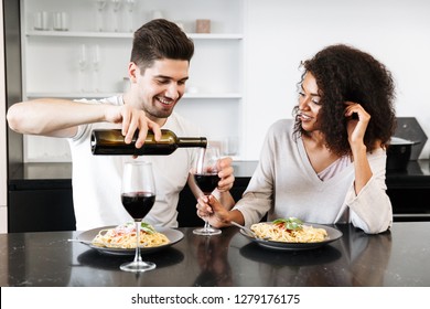 Beautiful Young Multiethnic Couple Having A Romantic Dinner At Home, Drinking Red Wine And Eating Pasta, Pouring