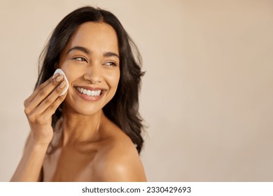 Beautiful young multiethnic woman with bare shoulder removing make up with cotton pad on cheek isolated against beige background. Smiling hispanic girl applying dry powder using cosmetic cushion.