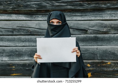 A beautiful, young, Muslim girl in a black veil with a closed face against a gray tree. In the hands of a sheet of paper. Copy space.