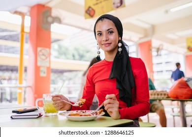 A Beautiful Young Muslim Malay Woman In A Hijab Head Scarf (tudung) Is Eating Her Breakfast Of Halal Nasi Lemak As She Sits In A Food Court During The Day. She Is Beautiful And Attractive. 