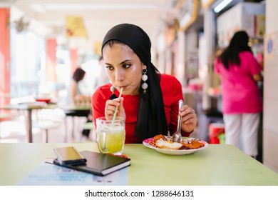 A Beautiful Young Muslim Malay Woman In A Hijab Head Scarf (tudung) Is Eating Her Breakfast Of Halal Nasi Lemak As She Sits In A Food Court During The Day. She Is Beautiful And Attractive. 