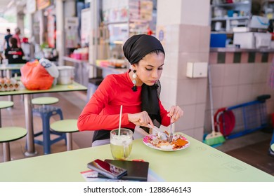 A Beautiful Young Muslim Malay Woman In A Hijab Head Scarf (tudung) Is Eating Her Breakfast Of Halal Nasi Lemak As She Sits In A Food Court During The Day. She Is Beautiful And Attractive. 