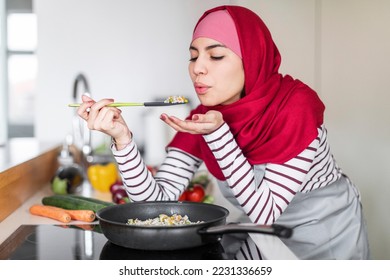 Beautiful young muslim woman in headscarf tasting meal while cooking dinner at home, standing next to electric stove with frying pan on and holding spoon, modern white kitchen interior, copy space - Powered by Shutterstock