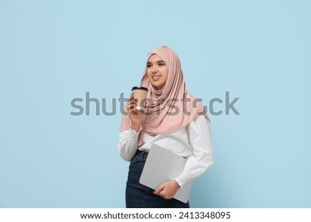 Beautiful young Muslim woman in hijab with paper cup of hot coffee and modern laptop on blue background