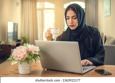 Beautiful young muslim woman wearing abaya drinking coffee and using laptop at home - Powered by Shutterstock