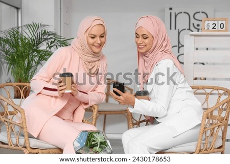 Beautiful young Muslim women in hijab with cups of coffee and mobile phone sitting at cafe