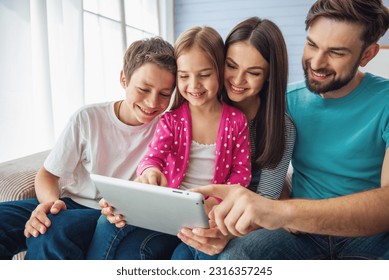 Beautiful young parents, their cute little daughter and son are using a tablet and smiling, sitting on sofa at home - Powered by Shutterstock