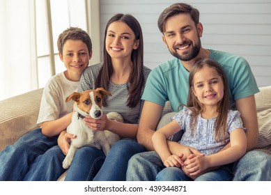 Beautiful young parents, their cute little daughter and son are looking at camera and smiling, sitting with their cute dog on sofa at home
