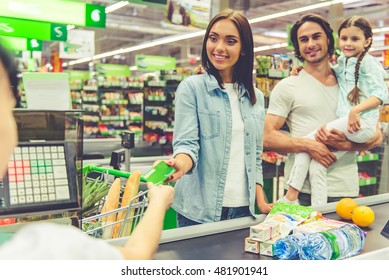 Beautiful young parents and their cute little daughter are smiling while standing near cash desk in the supermarket. Mom is giving a credit card - Powered by Shutterstock