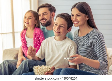 Beautiful Young Parents, Their  Daughter And Son Are Watching TV, Eating Popcorn And Smiling, Sitting On Sofa At Home. Mom Is Using A Remote Control