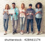 Beautiful young people of different nationalities are using gadgets and smiling, standing against white brick wall