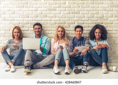 Beautiful young people of different nationalities are using gadgets, looking at camera and smiling, sitting against white brick wall