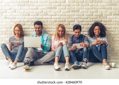Beautiful young people of different nationalities are using gadgets and smiling, sitting against white brick wall