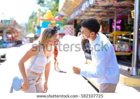 Beautiful young people, loving couple strolling in amusement park, looking at each other, kissing and cuddling, try candy floss, pose for camera and fooled at an amusement park outdoors. Girl with