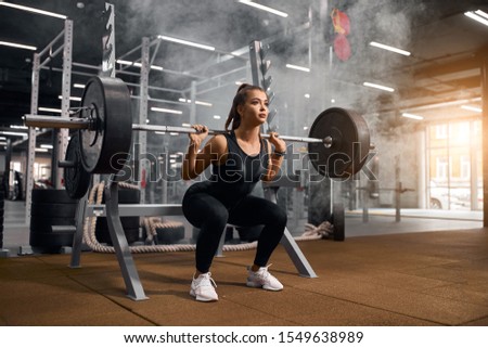 Beautiful young powerlifter squatting in modern fitness studio using heavy barbell, looking aside expressing self assurance, professional sport concept, white smoke in the air