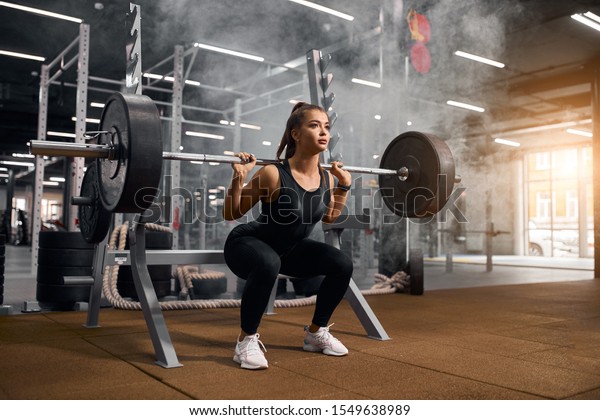 Beautiful young\
powerlifter squatting in modern fitness studio using heavy barbell,\
looking aside expressing self assurance, professional sport\
concept, white smoke in the\
air