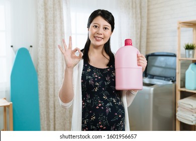 Beautiful Young Pregnant Asian Woman Is Holding Fabric Softener During Laundry Time. Elegant Smiling Future Motherhood Showing Ok Sign Looking At Camera. Happy Mom Holding Detergent Doing Housework.