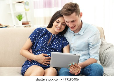 Beautiful Young Pregnant Couple With Tablet Computer Sitting On Sofa