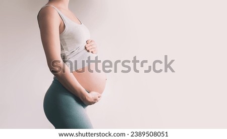 Beautiful young pregnant woman in good fit holds hands on her belly. Happy mother waiting for baby birth during pregnancy. Maternal health, maternity, preparation and expectation concept.