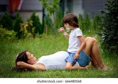 Beautiful Young Pregnant Woman And Her Older Child, Lying Down In The Grass, Making Soap Bubbles In Garden