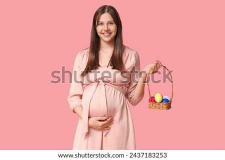 Beautiful young pregnant woman holding wicker basket with Easter eggs on pink background