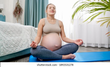 Beautiful Young Pregnant Woman In Leggings Meditating Against Bright Sun Shining Through Big Window At Bedroom. Concept Of Healthy Lifestyle, Healthcare And Sports During Pregnancy.