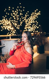 Beautiful young pregnant woman in red dress relaxing on her balcony in evening hours and drinking wine. 