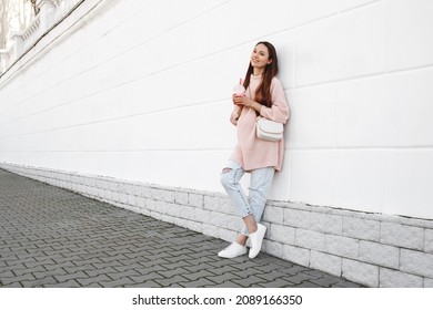 Beautiful Young Pregnant Woman Wearing Pink Sweater, Jeans And A Bag Standing Near White Wall On A City Street With A Drink In Her Hands.